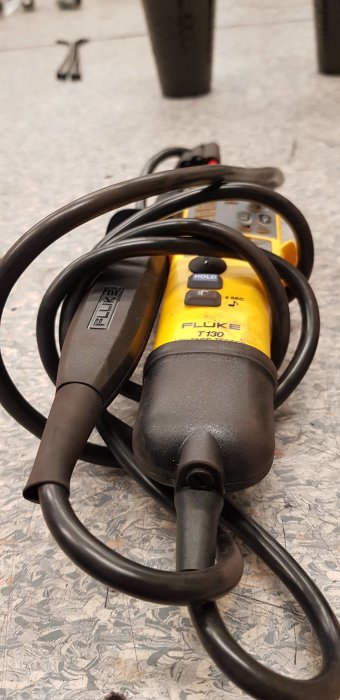 En multimeter med gul och svart design, märkt "FLUKE T130", och svarta testkablar mot en bakgrund av verkstadsgolv.