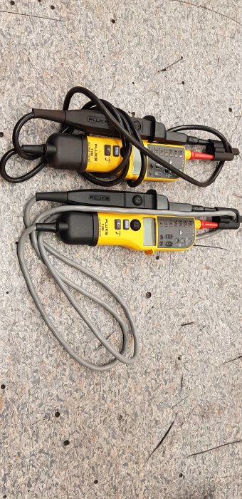 Två multimeters av samma modell, en med grå och en med svart kabel, på granitgolv.