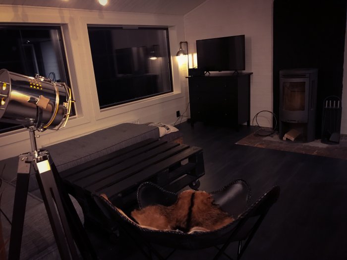 Mysigt vardagsrum med en fladdermusfåtölj i förgrunden, en TV, fönster och en braskamin i bakgrunden.
