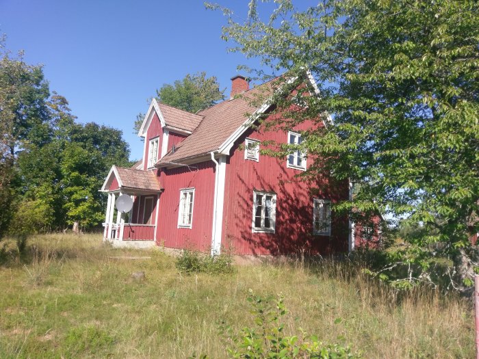 Rött traditionellt hus med vita knutar omgivet av grönska under en klarblå himmel.