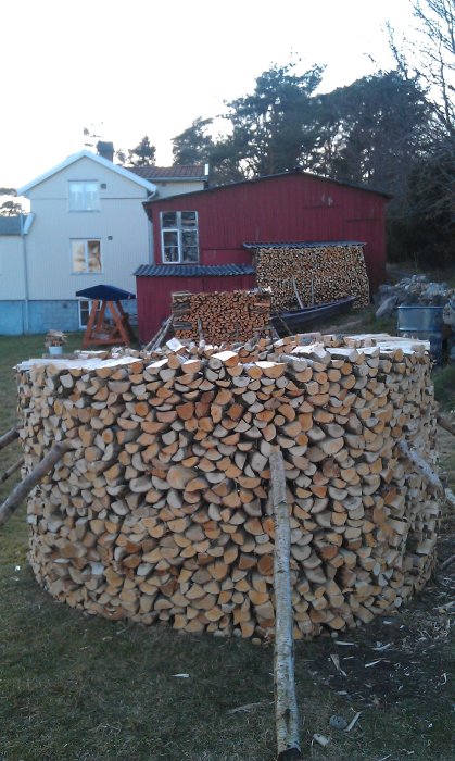 Stapel av ved med barken uppåt som ett tak, hus i bakgrunden.