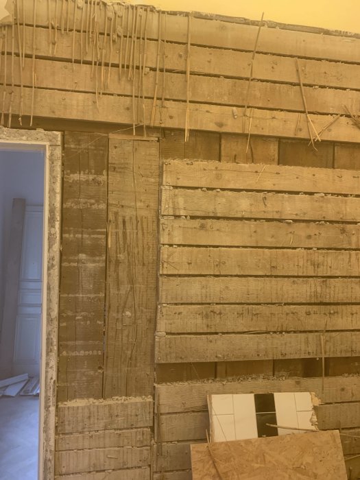 Seinä, joka on osittain purettu, paljastaen puurakenteita ja tiiliä, lähellä kynnystä vasemmalla. Yläosassa roikkuvia olkia tai säleitä.