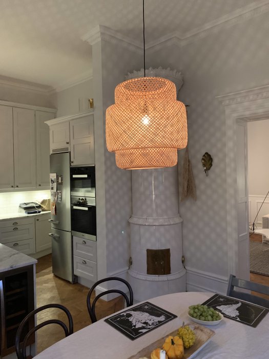 Valkoinen keittiö, jossa on takka ja roikkuva valaisin, pyöreä ruokapöytä edessä, hedelmiä ja lautasia pöydällä.