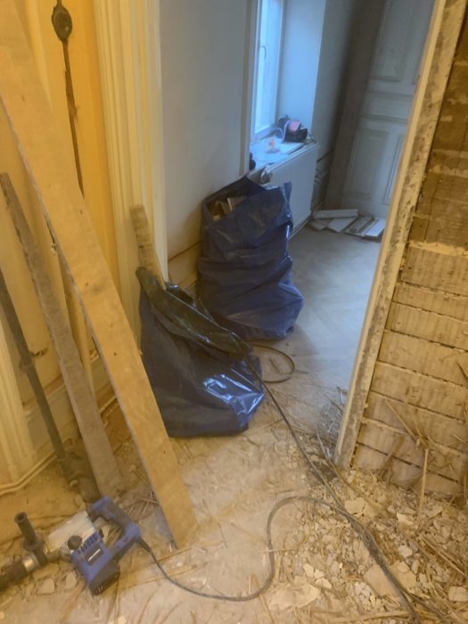 Avoin seinäaukko, lähellä sinisiä roskapusseja ja lattialla sähkötyökalu. Seinän materiaali näkyvissä, ympärillä rakennusjätettä.