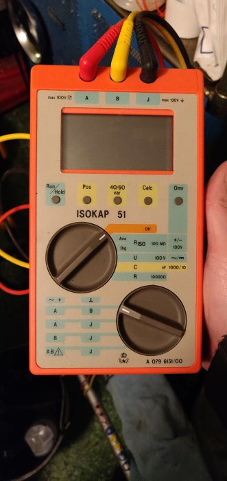 Orange ISOKAP 51 mätinstrument med testsladdar för elarbeten, hållen i handen framför en verktygsbakgrund.