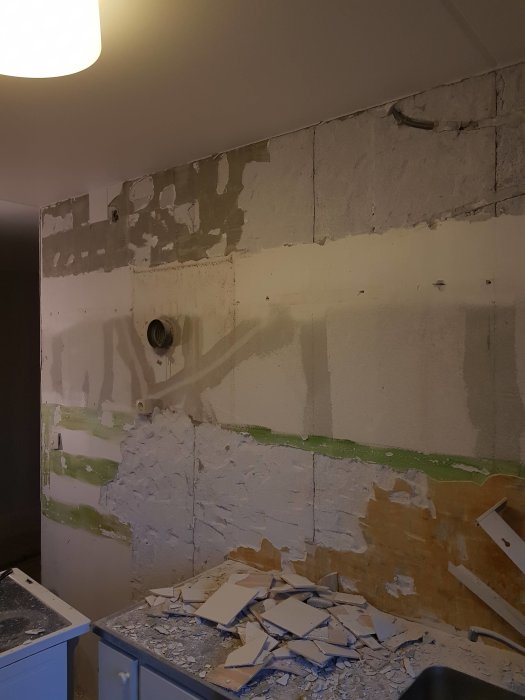 Renoverat kök med väggar delvis spacklade och borttaget kakel samt rör i väggen ovanför en köksbänk med skräp.
