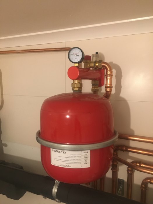 Röd expansionskärl med manometer för värmeinstallation, monterad på väggen med kopparledningar.