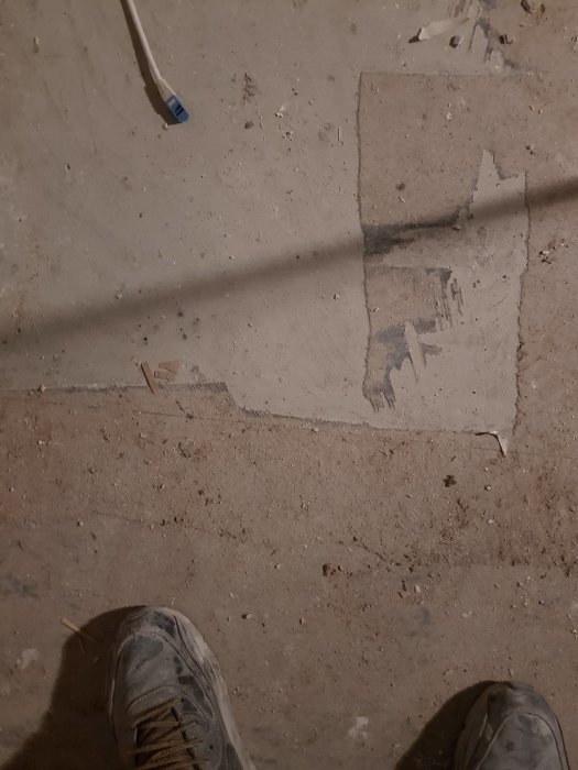 Betonggolv med delvis borttagen mattlim, skugga och synliga arbetsskor.