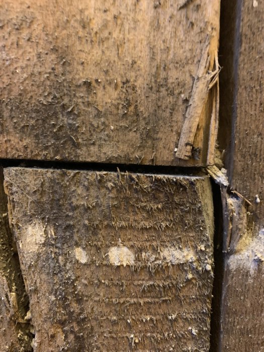 Lähikuva vanhan puupaneelin halkeamasta, jossa näkyy kulunut ja karheaksi purtunut pinta sekä pieni irronnut sälö.