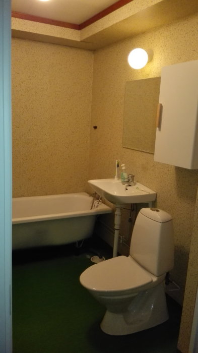 Gammaldags badrum med badkar, handfat, toalett och grönt golv i behov av renovering.