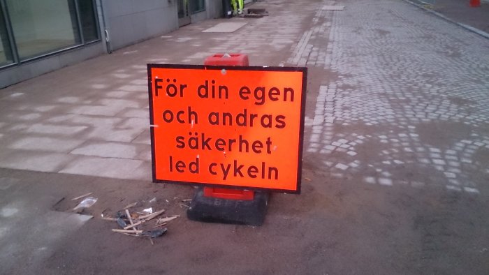 Orange skylt med texten "För din egen och andras säkerhet led cykeln" och spillror på marken.