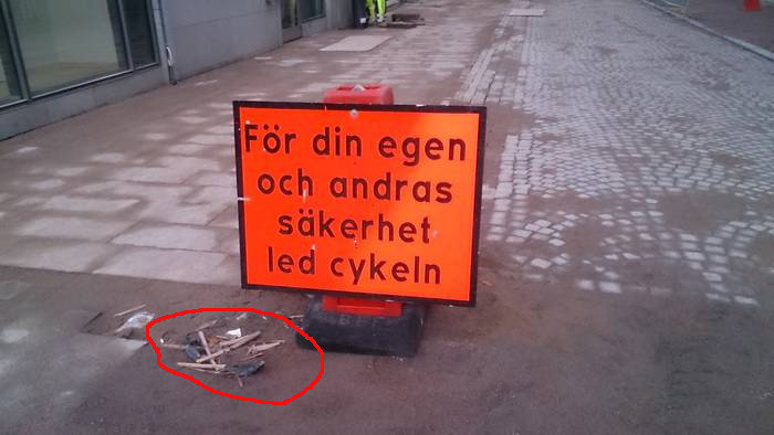 Delar av en förstörd cykel på marken bredvid en skylt som uppmanar "För din egen och andras säkerhet led cykeln