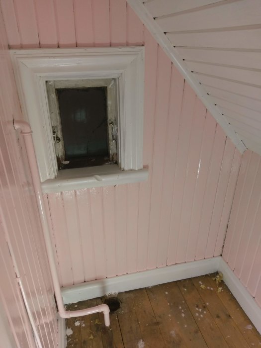 Hörn av ett rum under renovering med rosa väggar och en liten vitmålad garderobsdörr, samt synlig golvrörledning och trägolv.