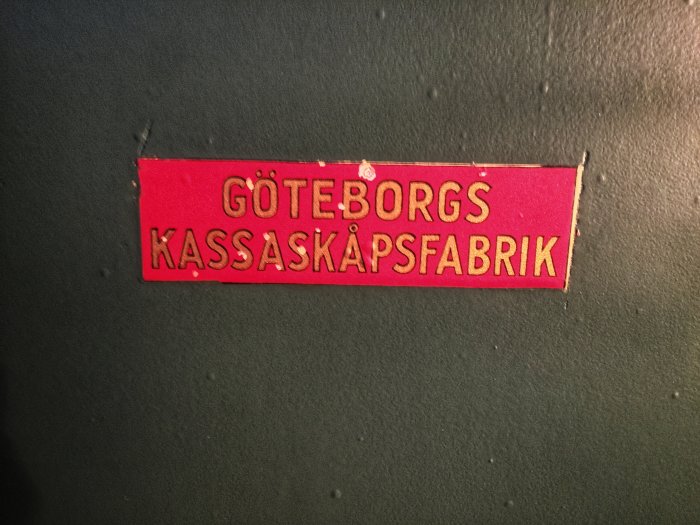 Närbild på en sliten etikett med texten "GÖTEBORGS KASSASKÅPSFABRIK" på en grönmålad yta.