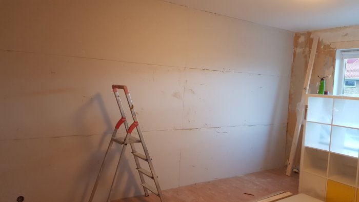 Renoveringsarbete med bortriven tapet och limmad ytgipsskiva på en vägg i en lägenhet, stege i förgrunden.