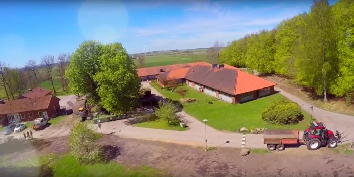 Flygbild över en skola med röda tak och en traktor utanför, omgiven av gröna träd och fält.