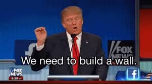 Person som gestikulerar under en debatt med texten "We need to build a wall." på skärmen.