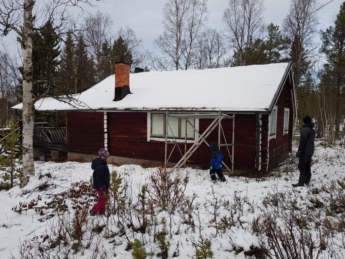 Två personer betraktar en röd stuga med snötäckt tak och altan i vinterskog.