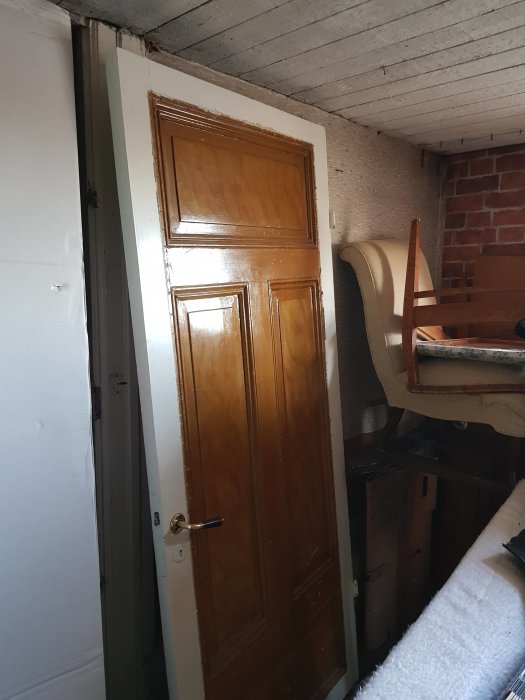 Brun dörr med paneler och dörrhandtag, står lutad mot vägg i rörigt utrymme.