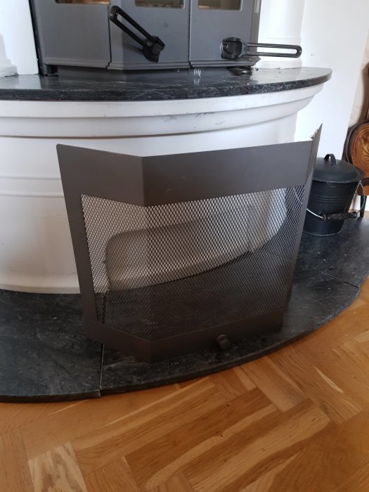Nyskick gnistskydd i metall för Handöl braskassett står på ett marmorgolv framför en vedspis.