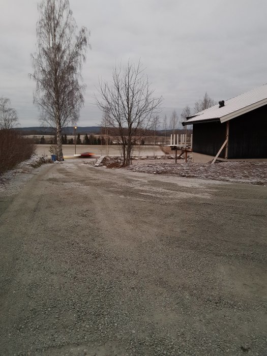 Nyligen grusad gårdsplan med jämnt fördelat grus framför en byggnad, med träd och grå himmel i bakgrunden.