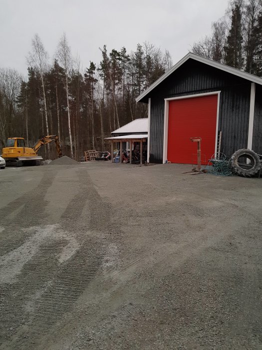 Nygrusad gårdsplan framför röd lada med vit snö på taket, grävmaskin i bakgrunden.