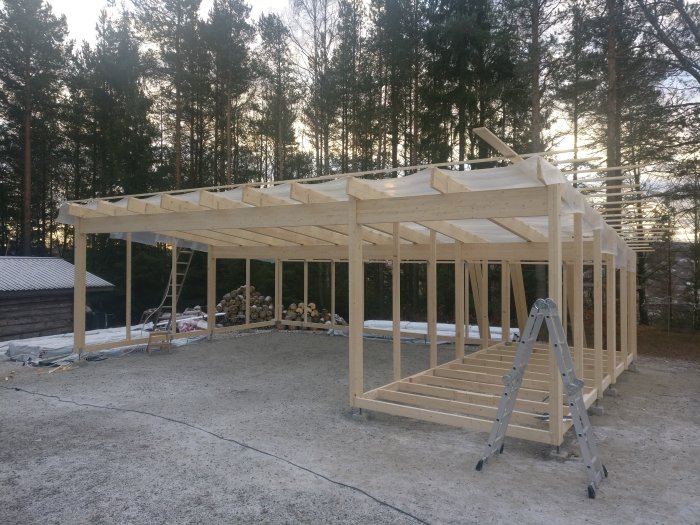 Byggnation av carport med trästomme och takbjälkar, delvis täckt tak och stege i förgrunden.