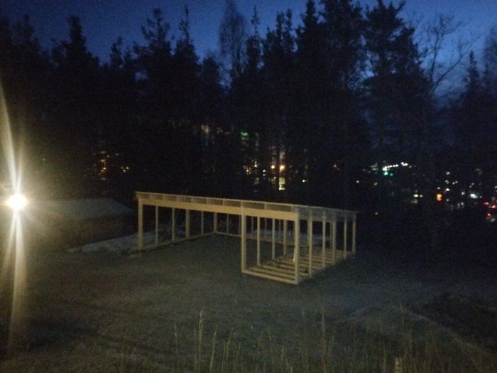 Delvis konstruerad carport med takbjälkar i skymningen, belysning och träd i bakgrunden.