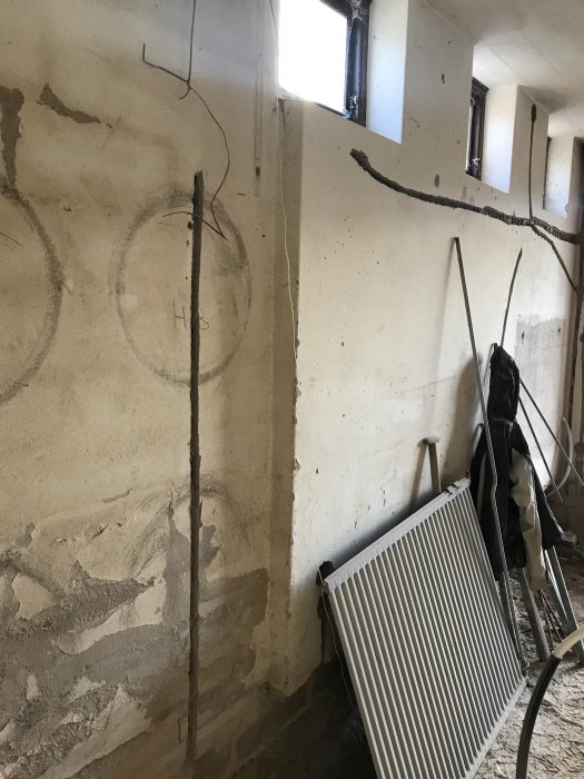 Renoveringsarbete med synlig påbyggd vägg och rördragningar, bredvid en liggande radiator, osorterade objekt i rummet.