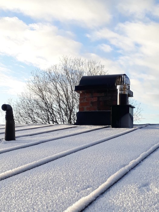 Vindflöjel och snöbelagd takplåt på ett platt tak med tegelskorsten i bakgrunden.