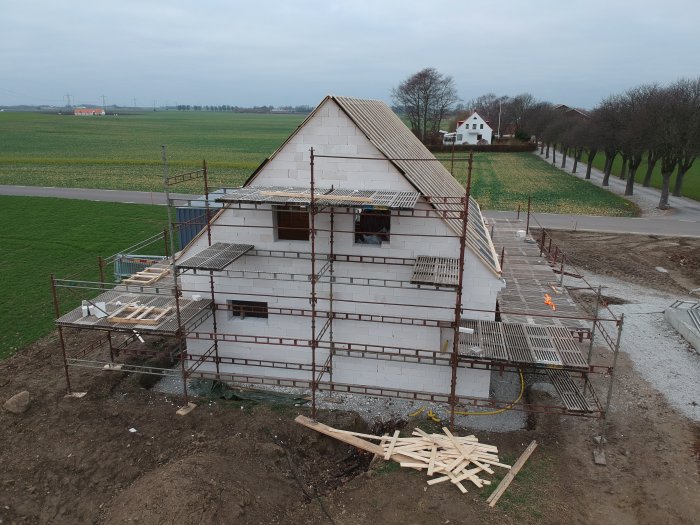 Byggarbetsplats med ställningar runt en halvfärdig gavel av ett hus, material på marken, mot öppen landsbygd.