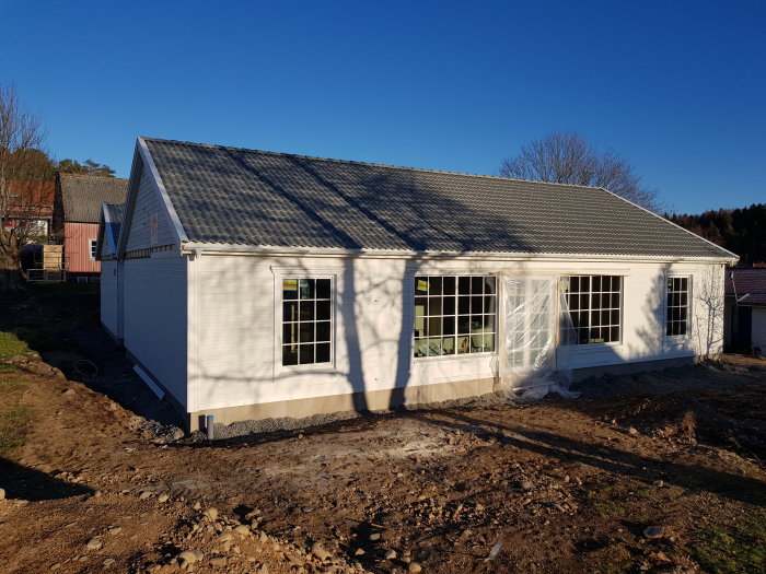 Nytt hus med vit fasad och grått tak i soligt väder, byggarbetsplats med jord och grus framför huset.