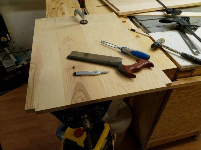 Verktyg och trämaterial på en arbetsbänk, inklusive handsåg, stämjärn och hammare, med pågående snickeriprojekt.
