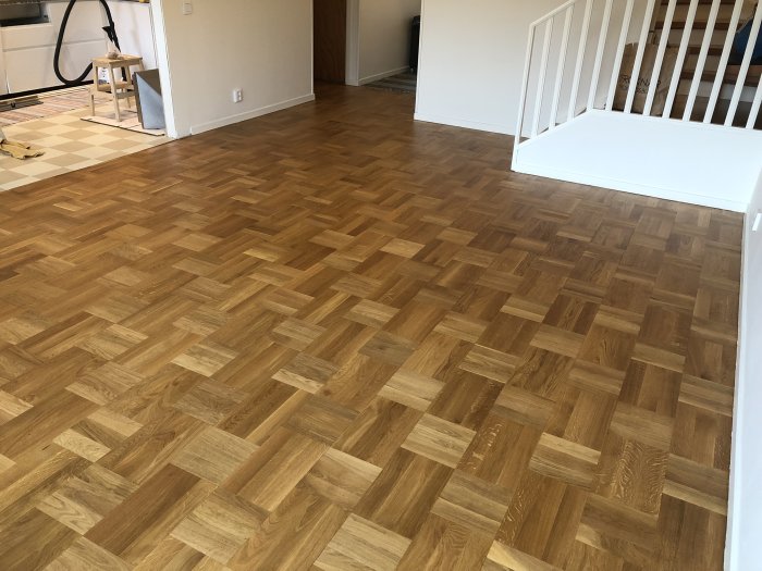 Nyinstallerat parkettgolv i anslutning till rum med rutigt linoleumgolv och vit trappa.
