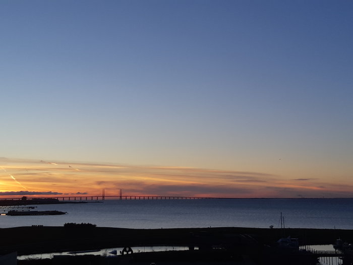 Solnedgång med orange himmel över en silhuett av en lång bro över vatten.