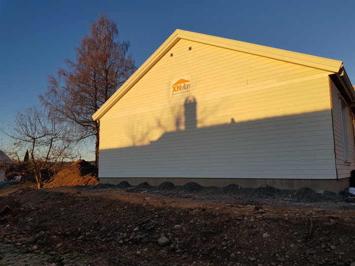 Grävarbete runt hus med nytt grus och jordhögar i solnedgång med skuggor på vit fasad.