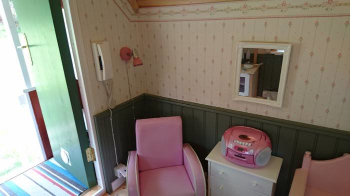 Rosa gömd väggtelefon vid öppen dörr i ett rum med blommig tapet, grön panel och inredning i pastellfärger.
