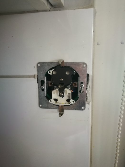 Trasigt elektriskt vägguttag utan frontpanel monterat på en vit vägg med synliga kablar och komponenter.