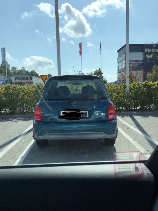 Blå Nissan Micra parkerad utomhus på en solig dag, sett från en annan bils förarsida.