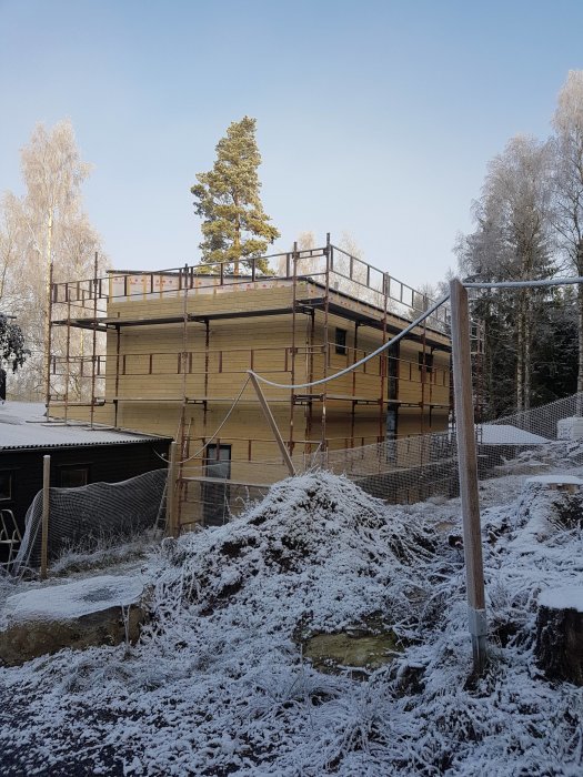 Hus under renovering med snötäckt träpanel och byggnadsställningar runt omkring i vinterlandskap.