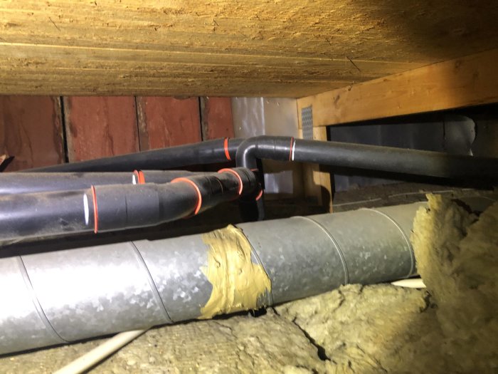 Oisolerade rör och ventilationskanaler i ett trångt utrymme under takbjälkar.