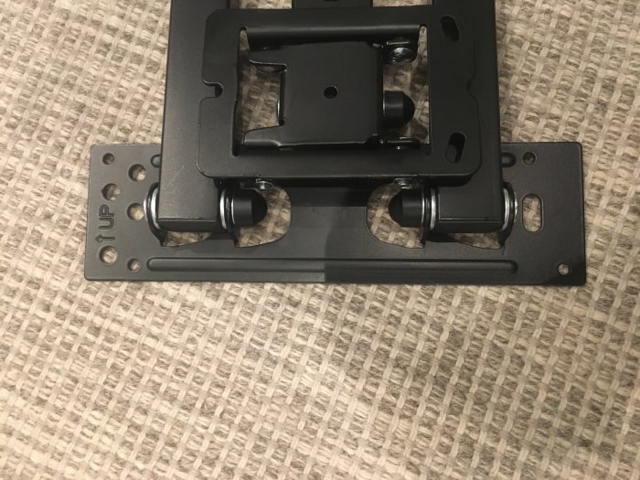 Ett svart fäste med fyra mindre och två större hål på en textilbakgrund, 24 x 7 cm storlek.