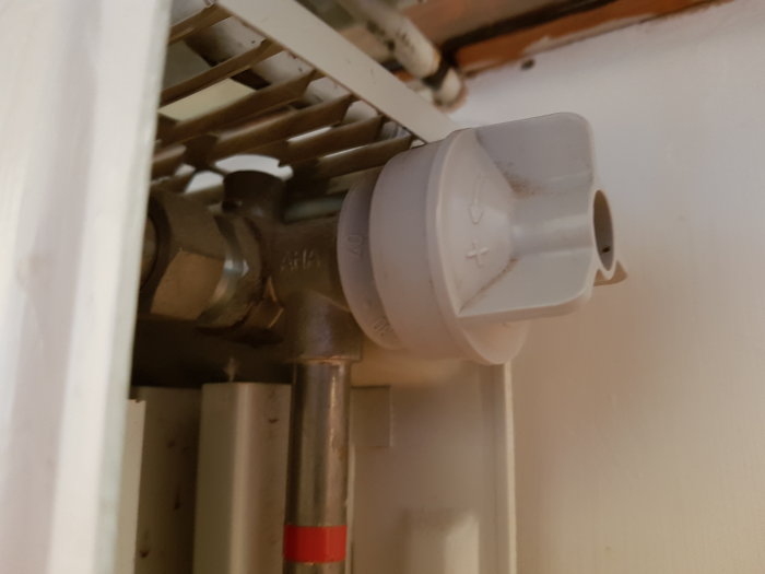 Äldre vit radiatorventil och termostat fastsatt vid en värmeelement med synliga rör och en vägg i bakgrunden.