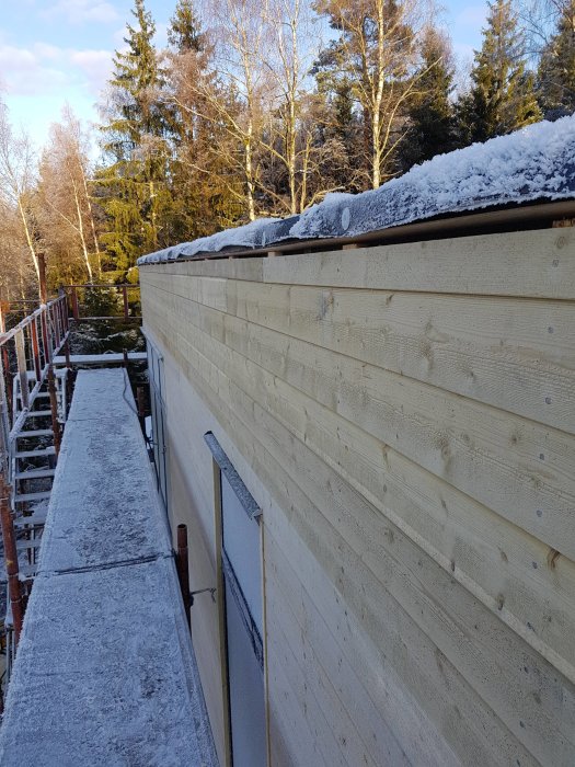 Nyuppsatt träpanel på husvägg med snötäckt takkant i soligt landskap.