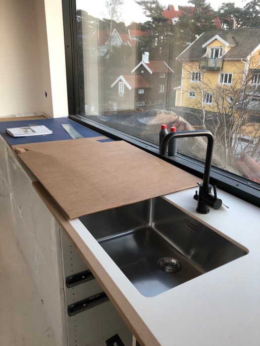 Nyinstallerad köksbänk med ränna och svart kran framför ett fönster, skyddad med brunt papper.