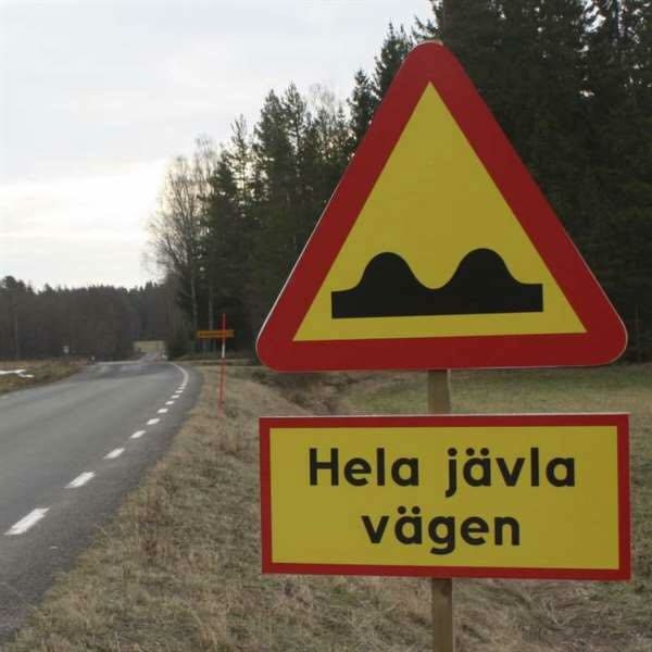 Varningsskylt med ojämn väg-symbol över humoristisk skylt som säger "Hela jävla vägen" vid sidan av landsväg.