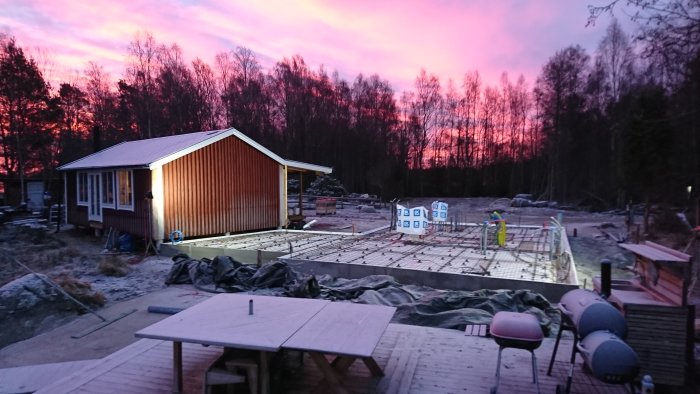 Betongplatta i gryningen med färdig armering nära skogsbryn och hus, himmel i rosaskimrande färger.