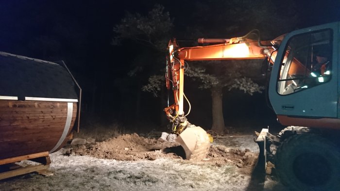 Grävmaskin i arbete på kvällen, belyser en skogsmark och gräver i jorden nära en träkonstruktion.