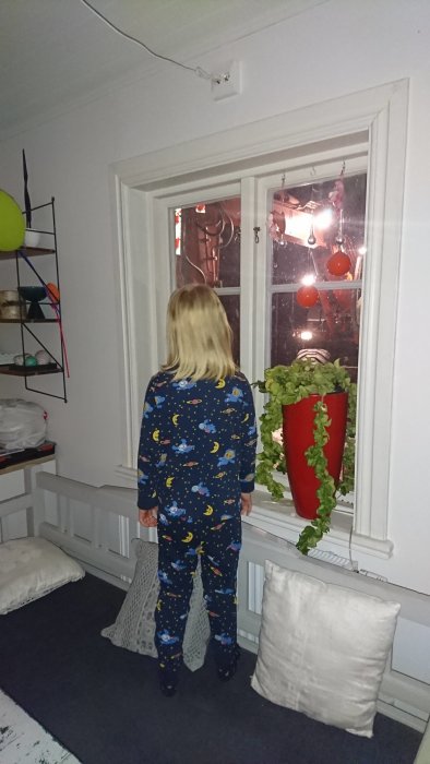 Barn i pyjamas med rymdmönster tittar ut genom fönster på en grävmaskin på kvällen.