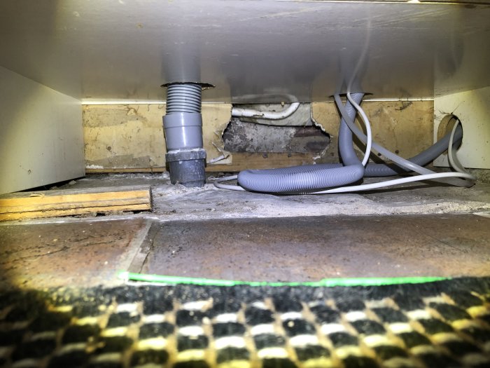 Synligt hål vid golvregel under köksskåp, glipor och rostiga skruvar som indikerar fuktproblem.